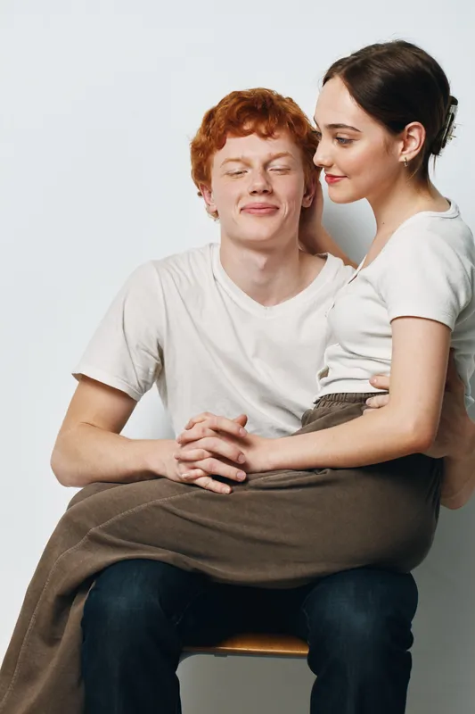 Couple sitting together in casual clothes with affectionate expression and closed eyes on white background. Young man and woman sharing intimate moment, comfort and closeness.