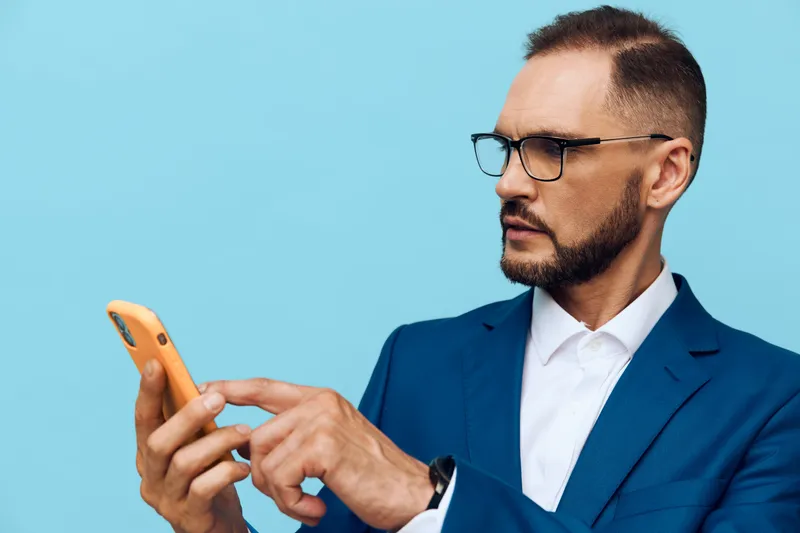 Professional businessman stands against a solid blue background, focused and thoughtful while using a smartphone. Isolated color backdrop emphasizes corporate style, calm mood, and confident demeanor.