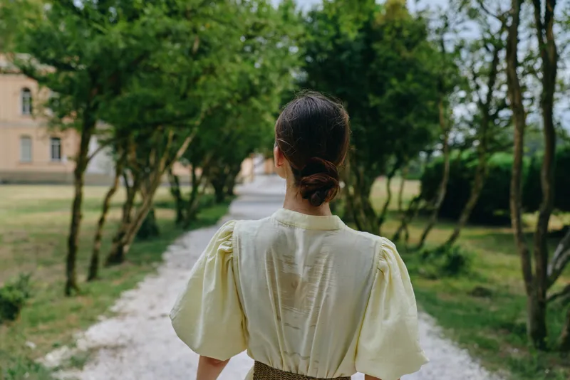 Woman seen from behind walking along a tree lined path in a sunny park, wearing a pale yellow blouse, creating a peaceful outdoor scene for photography
