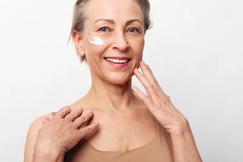 Smiling mature woman with skincare cream on cheek applies moisturizer to face, showing natural beauty and wellness. Studio portrait on white background with soft lighting for skincare concept.