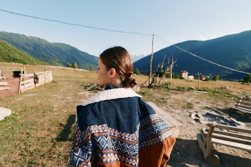 Girl in patterned sweater back view in rural village landscape with wooden fences and distant mountains, outdoor portrait of youth exploring countryside pathway under clear blue sunlight.
