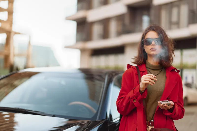 A woman in a red coat smokes beside a sleek car and checks her phone in a modern urban setting, conveying a candid city lifestyle moment.
