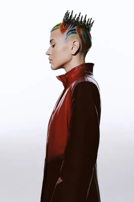 Woman with colorful mohawk hairstyle and crown, wearing a red leather jacket, standing in profile against a white background, displaying confident edgy style.