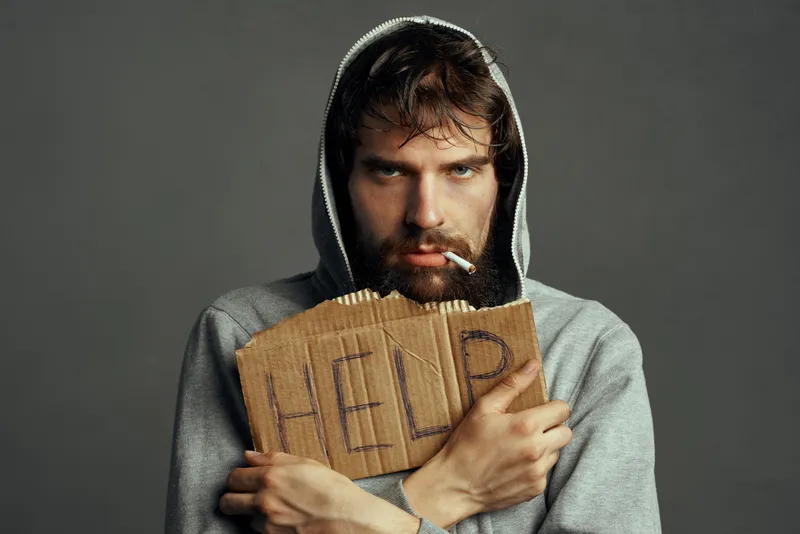 Бородатый мужчина в капюшоне с сигаретой держит картонку с надписью help