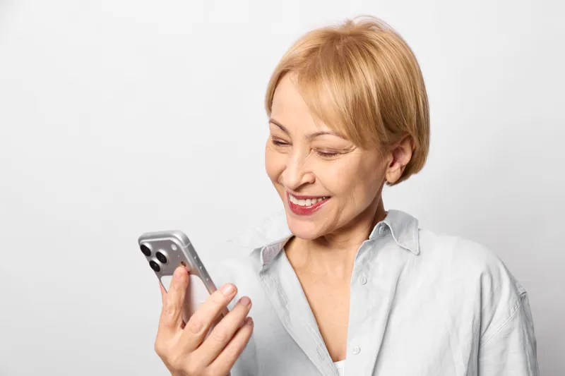 Senior woman smiling using smartphone, casual portrait, typing message, friendly communication mood, isolated on light neutral background, closeup, calm