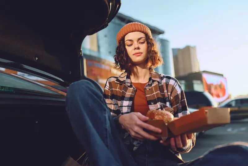Young woman eating burger in car trunk in urban setting, candid moment with authenticity and golden hour glow, mindful living vibe and emotional storytelling in relaxed portrait.