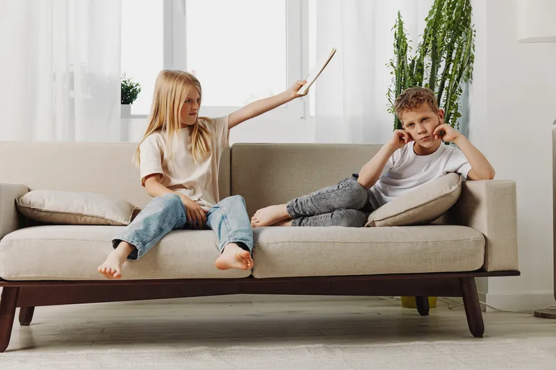 Two children sit on beige sofa in bright living room, girl playfully holds wooden stick near boy covering ears, family interaction concept.