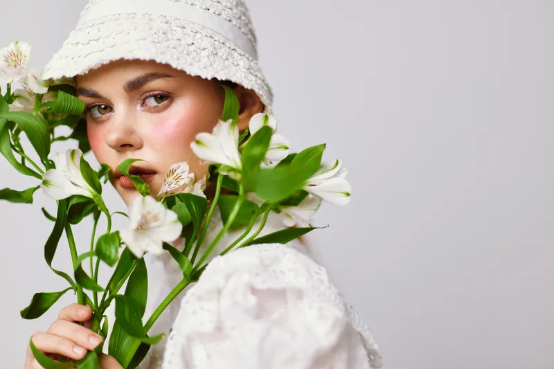 Young woman with a floral bouquet, wearing a white hat and a stylish outfit, expressing a serene emotion against a minimalistic background