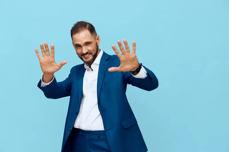 A confident business professional stands on a solid colored background, presenting an expressive pose with hands raised, conveying openness, energy, and approachability.