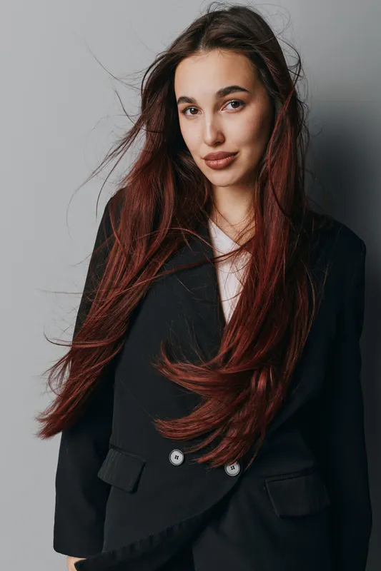 Young woman with long brown hair wearing black blazer posing against gray background. Portrait of confident female model with natural makeup and casual style for fashion or beauty concept.