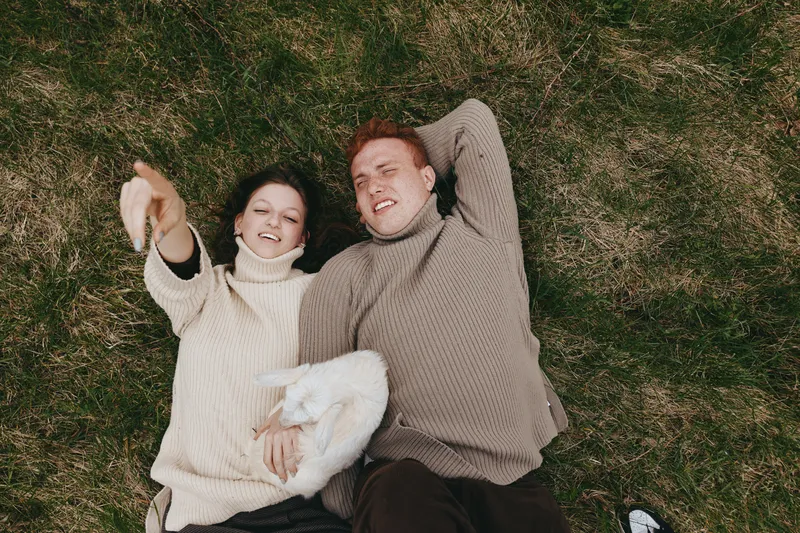 Couple lifestyle outdoor, lying on grass, smiling with eyes closed, cozy sweaters, sunlit moment, gentle breeze, natural colors, playful goat kid beside them, relaxed mood, candid connection and