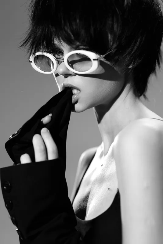 Black and white portrait of a woman wearing white sunglasses and biting sleeve with a stylish edgy mood.
