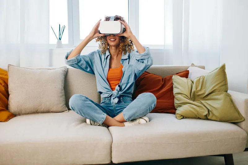 Улыбающаяся женщина наслаждается футуристической VR-игрой дома, в VR-очках, сидя на диване в современной гостиной