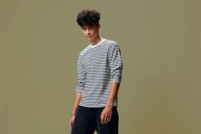 young man wearing striped shirt and black pants stands confidently against plain olive background, casual style and relaxed posture, studio portrait