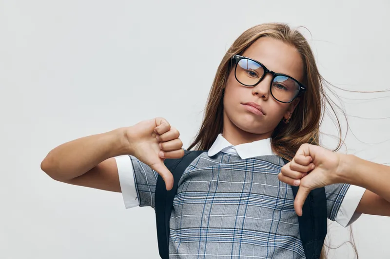 Girl with glasses and long hair wearing checked dress and backpack showing double thumbs down gesture against white background. Portrait of unhappy schoolgirl expressing disapproval.