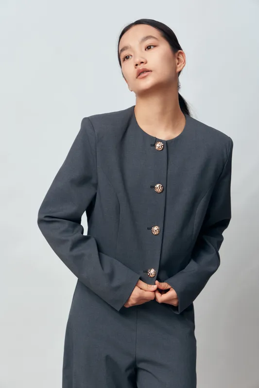 Young woman in gray business suit standing against light background looking away with confident thoughtful expression and hands near jacket buttons indoors studio portrait.