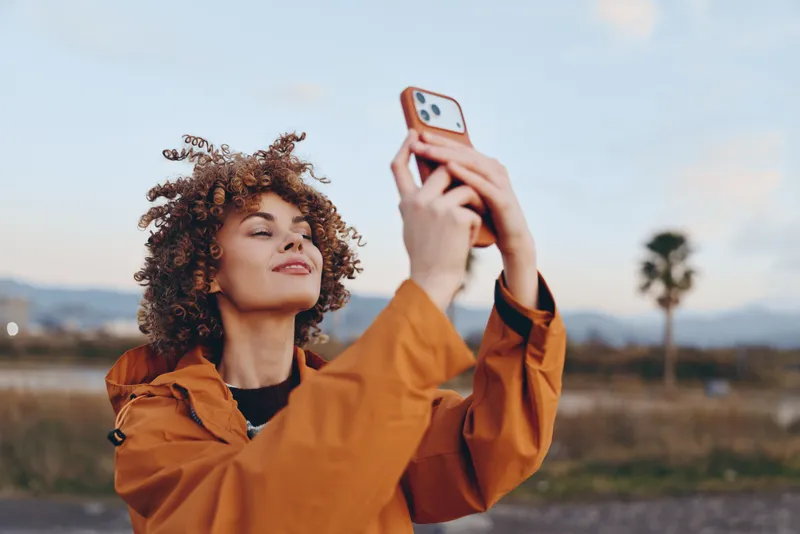 Woman lifestyle outdoors, curly haired woman in orange jacket holds smartphone for selfie, gazing ahead in warm daylight, blurred nature background, relaxed travel mood.