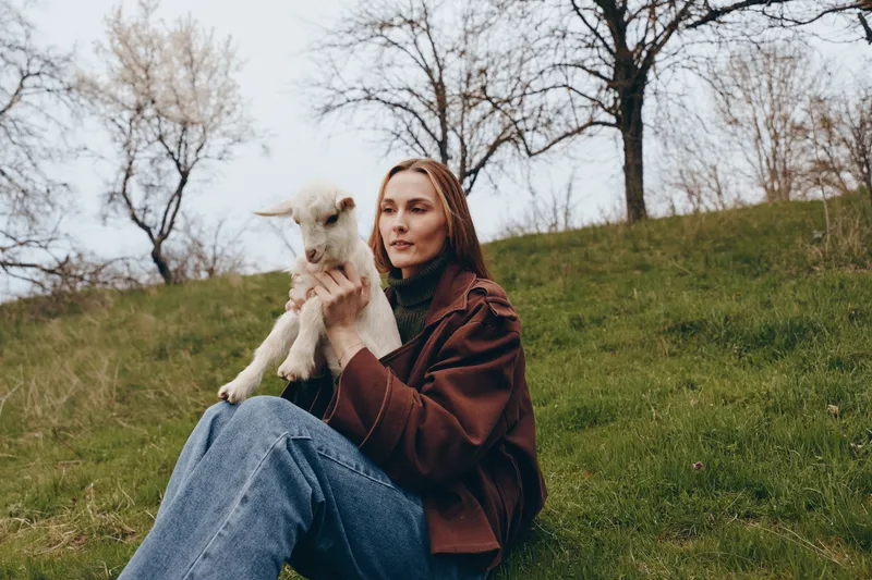 A woman in a brown coat sits on a grassy hill holding a young goat, outdoors. Warm light and a tranquil expression capture a lifestyle moment of connection between person and animal in nature.