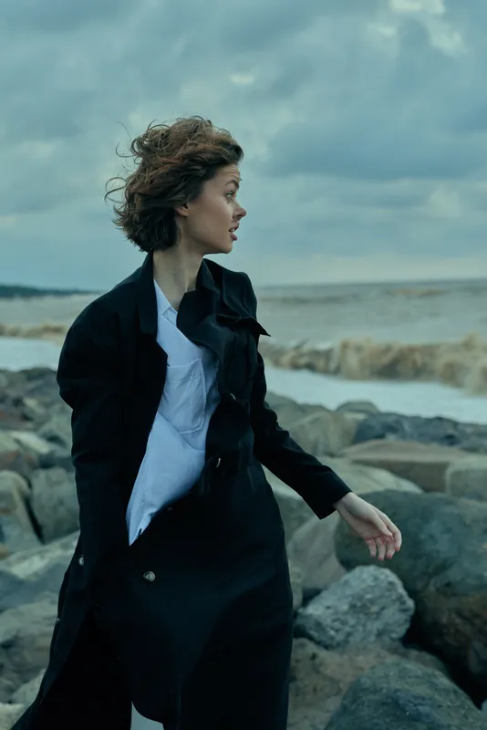 Woman walking along rugged coast in windy weather, wearing a dark coat and white shirt, flowing hair, sea in the background, moody atmosphere and motion