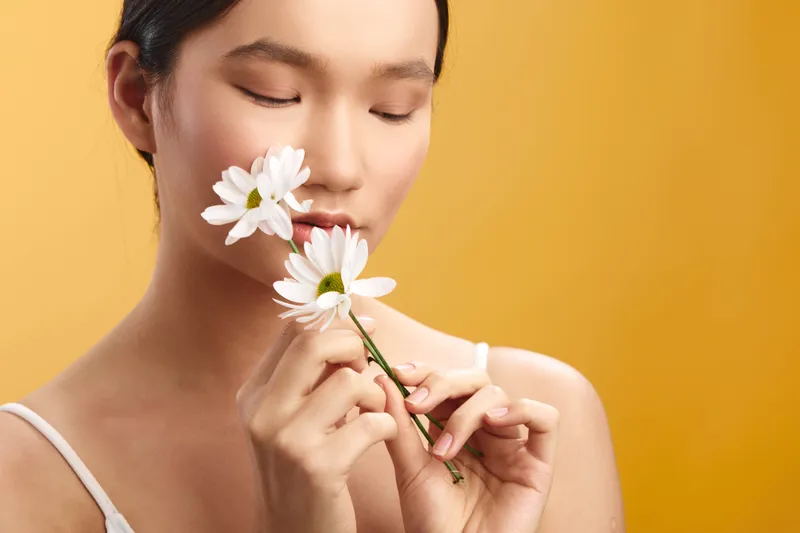 asian woman gently smelling daisies in a soft studio portrait. She closes her eyes with a serene smile, evoking calmness, wellness, and natural beauty, reflecting inner peace for lifestyle imagery.