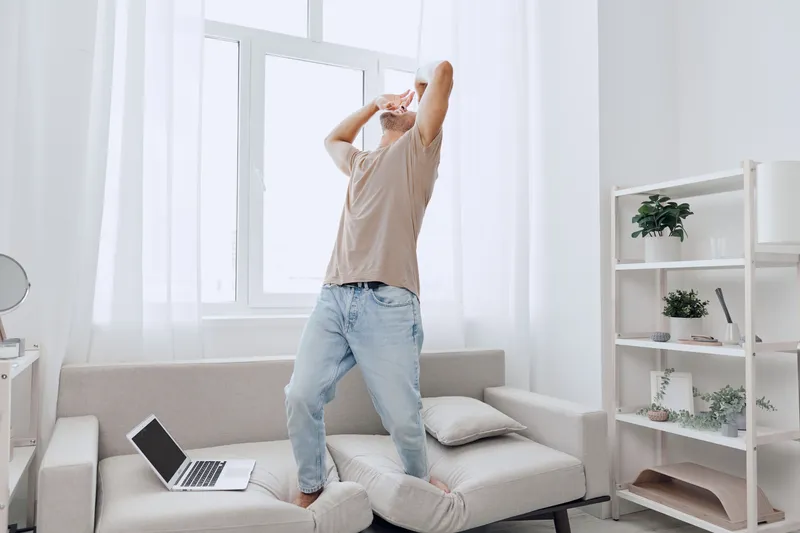 Joyful man enjoying a refreshing drink while standing on the couch in a minimalistic modern living room. Bright, airy space with natural light streaming in.