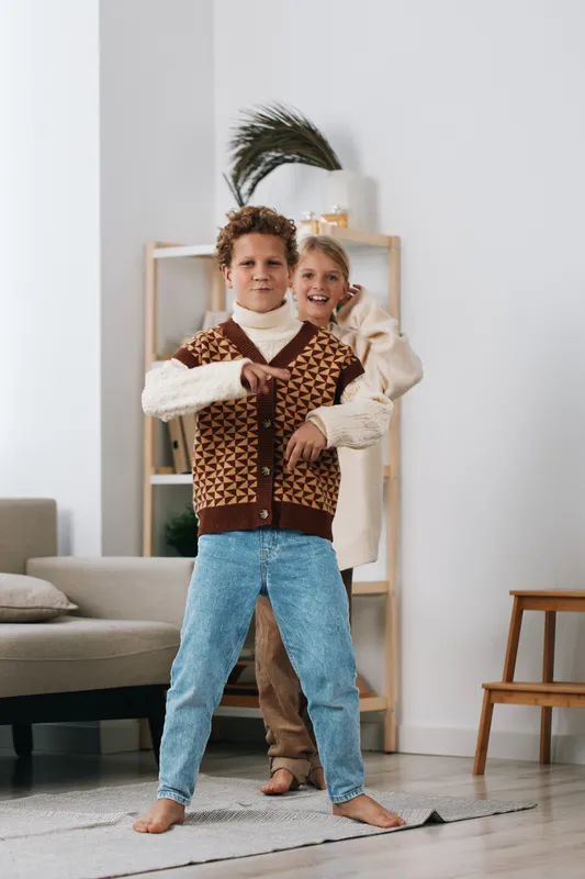 Two playful children dance indoors, smiling and energetic, wearing retro knit vest and denim pants, in a bright living space with soft furnishings and a calm, welcoming atmosphere.