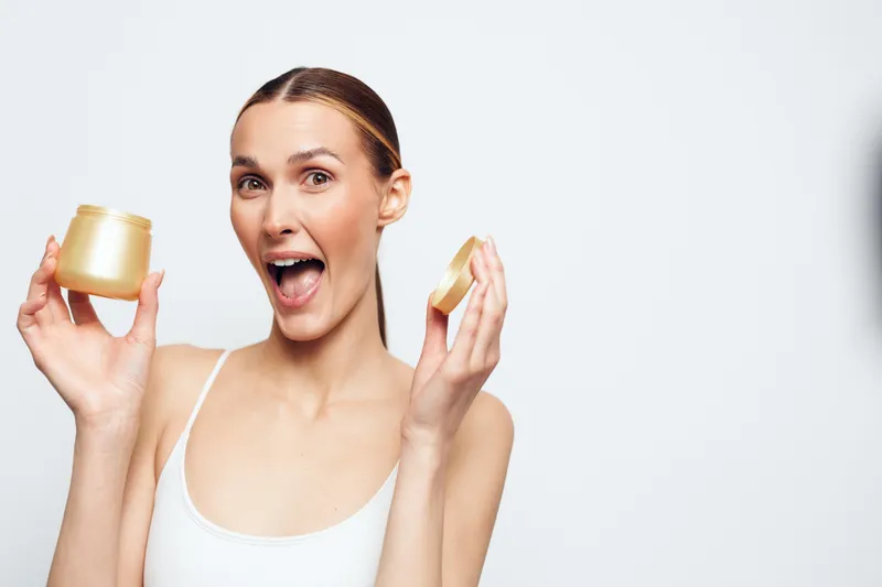 A smiling young woman with brunette hair holds two gold cosmetic jars in front of a bright white studio backdrop, showcasing skincare products and excitement.