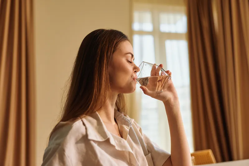 Woman drinking water from glass at home in morning, relaxed portrait by window with curtains, casual shirt, healthy hydration routine and calm indoor lifestyle scene
