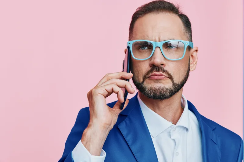 A business professional man speaks on a phone against a solid pink background, projecting confidence, focus, and a polished corporate demeanor for an executive lifestyle shot.