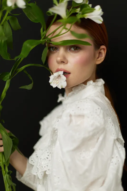 Young woman with flower in mouth, wearing white blouse, against dark background, conveying emotions of serenity and innocence in a conceptual portrait