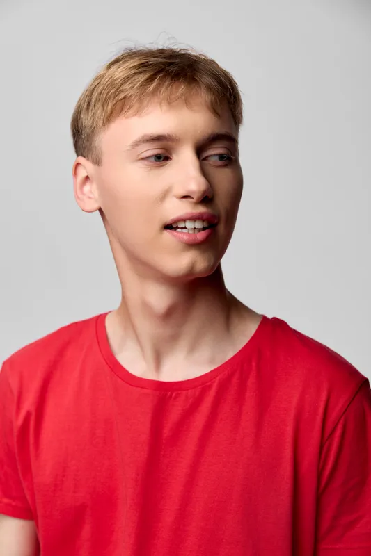 Young man in a red shirt turns slightly away, casual style and relaxed expression, studio lighting highlights clear skin and soft shadows, minimal gray backdrop, modern fashion photography vibe