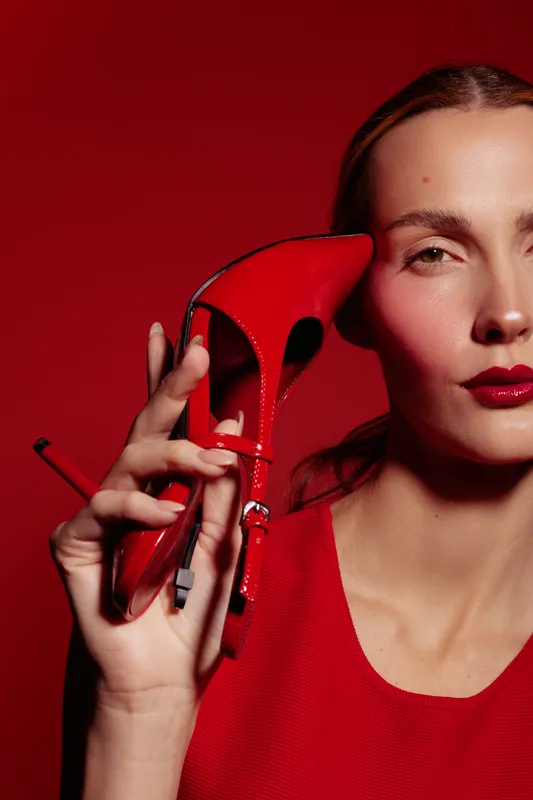 Close-up of a woman in red dress holding a red high heel shoe near her face against a red background. Concept of fashion, style, elegance, and bold color contrast.