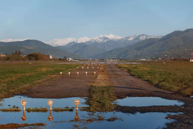 Abandoned rural runway with puddles reflecting runway lights stretches toward snow-capped mountains under a clear blue sky in a peaceful natural landscape.
