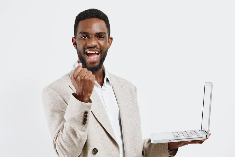 Joyful young African American man in beige blazer holding a laptop computer, smiling confidently, expressing success and excitement, in a studio setting with plain white background. People lifestyle c