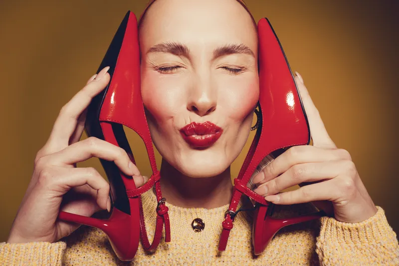 Happy woman with closed eyes holds red high heels near face, wearing yellow sweater. Concept of fashion, style, beauty, and joyful emotions in studio with warm background lighting.