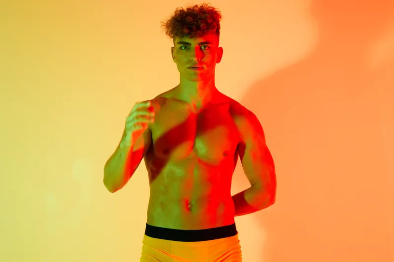 Young muscular man posing shirtless with colorful red and green lighting on yellow background, fitness model showing toned body and confident expression.