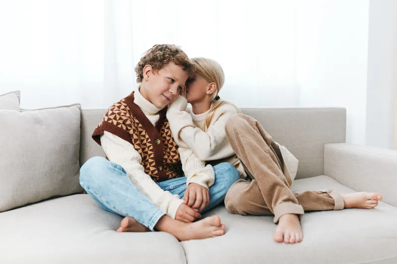 A couple sits close on a light sofa, wearing cozy knit sweaters, sharing a tender moment of warmth and comfort in a bright living room.