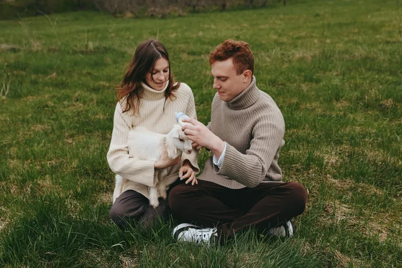Young couple sitting on grass in cozy sweaters gently holding a baby goat while enjoying a calm autumn day outdoors in a natural lifestyle setting.