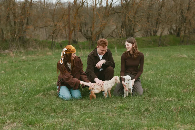 Casual outdoor scene with three people and goat kids in a grassy field, capturing joyful interaction, gentle touch, and lifestyle. Natural light highlights warmth, friendship, and animal