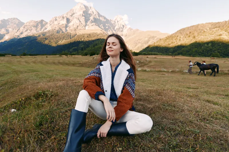 Woman sitting on a meadow with closed eyes in a knit sweater and boots, enjoying mountain scenery and nature air; peaceful relaxation with distant horse and rider in countryside landscape.