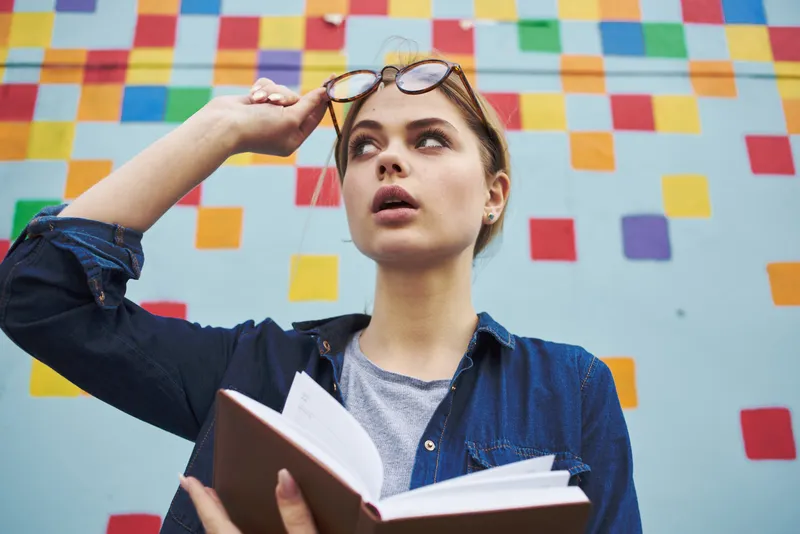 Студентка в рубашке и очках держит в руках книгу на улице возле граффити на стене