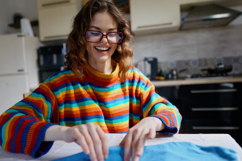 A cheerful woman in a bright striped sweater sits at a kitchen table, focused on sewing fabric. A cozy home lifestyle scene highlighting creativity, hobbies, and domestic tasks.