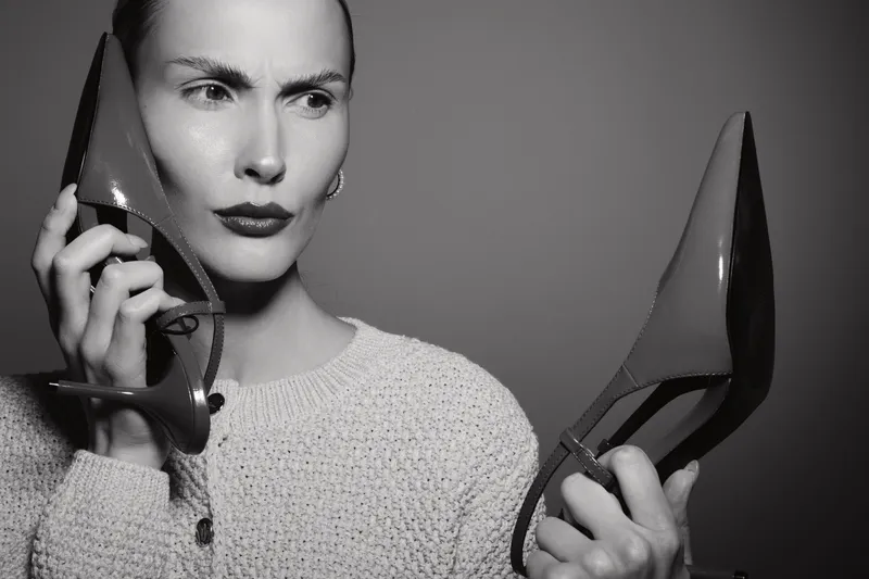 Serious white woman with pale skin examines two high heel shoes holding them close to face indoors. Concept for fashion choice and style decision.