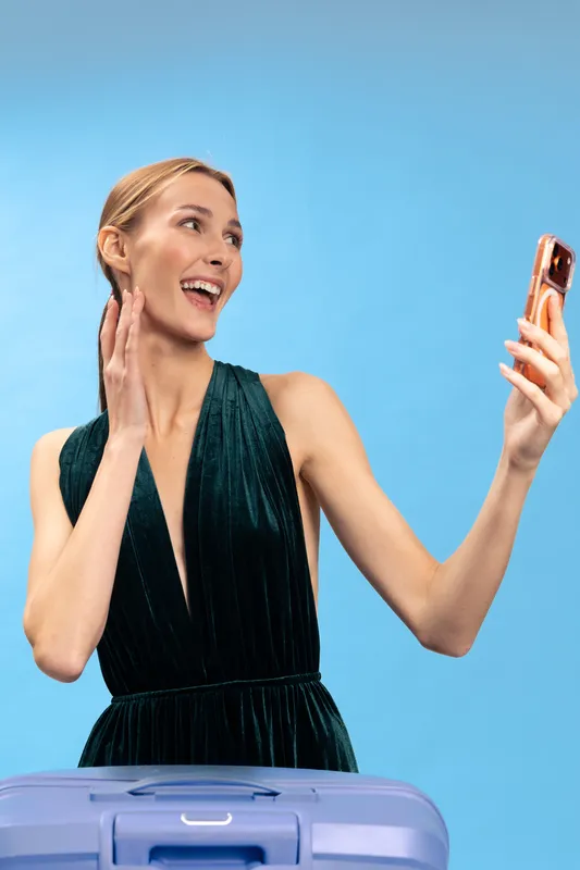 Smiling woman in elegant dark green dress takes selfie with smartphone, posing near purple suitcase on blue background. Concept of travel, fashion, and joyful vacation moments.