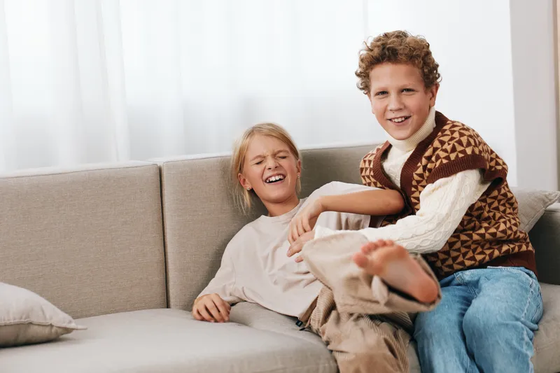 Two cheerful children relax on a soft sofa indoors, smiling and laughing as they share a playful moment together, warm natural light filling the cozy room.