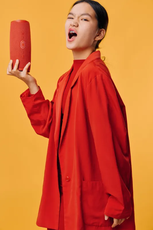 Woman with long black hair wearing oversized red jacket and holding red clutch purse on orange background. Fashion model posing with intense expression in studio lighting.