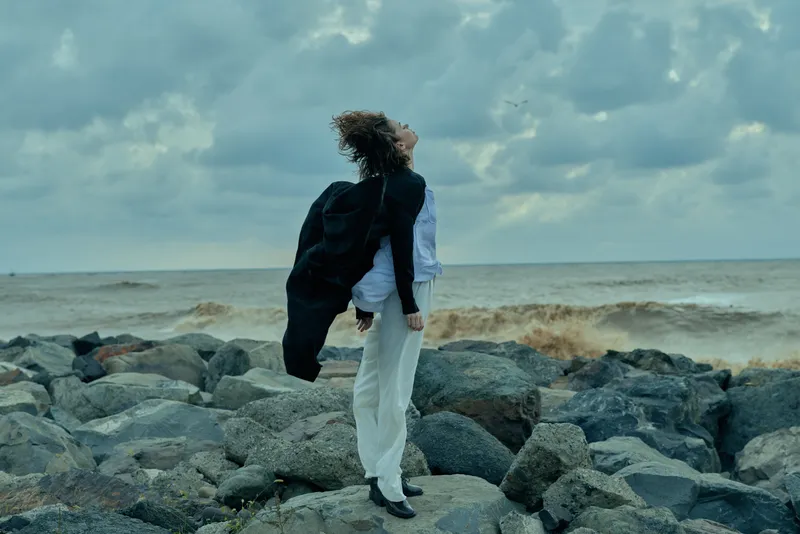 Windblown woman stands on rocky coastline by the sea, wearing a draped coat, looking toward the horizon under dramatic clouds, conveying resilience, solitude and coastal fashion
