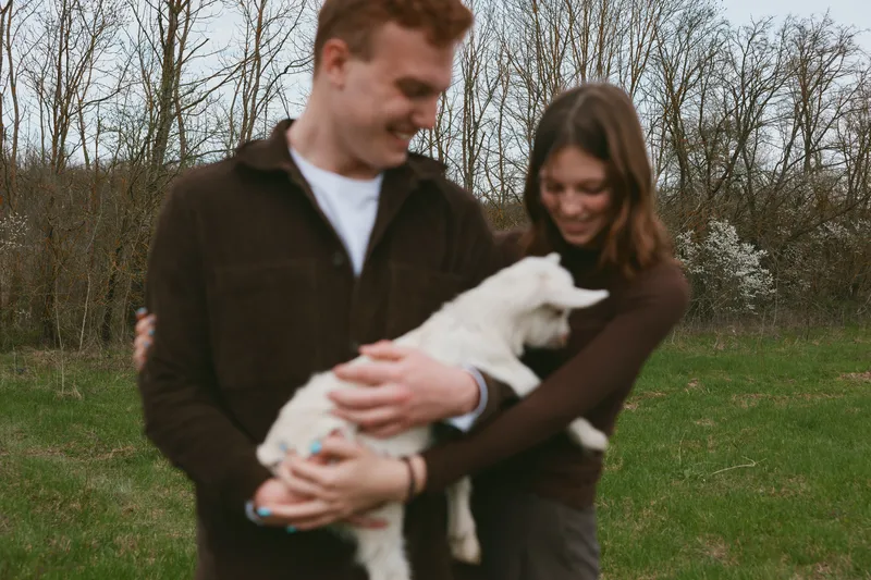 Happy couple outdoors with a playful goat kid, they cradle the young animal and share a warm smile, a candid lifestyle moment set in a grassy rural yard, peaceful and joyful