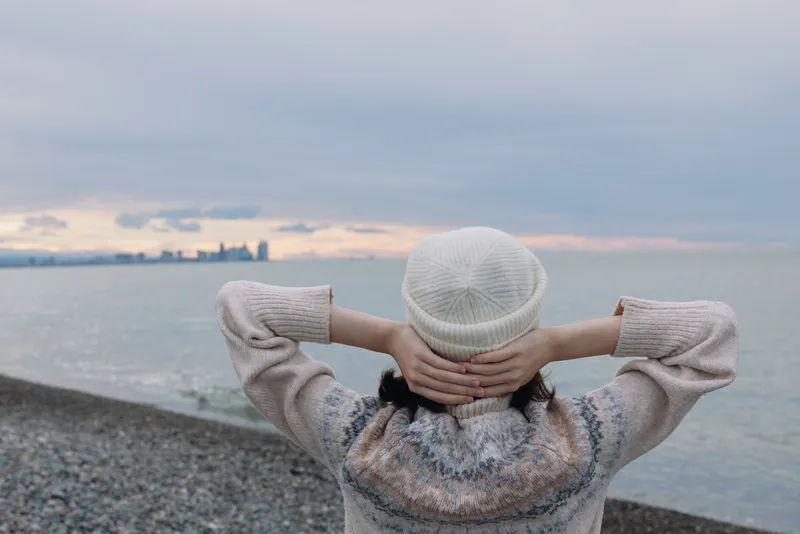 Contemplative person on a pebbled beach with hands behind head, wearing a knit sweater and beanie, gazing at calm water and distant city skyline at dusk.