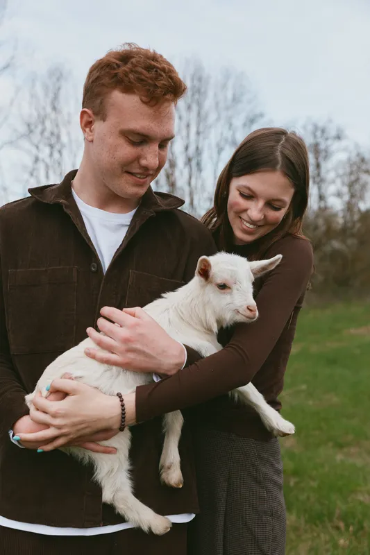 Joyful couple cradle a white goat kid in a sunlit field, showcasing tender bonding and rustic charm during a casual countryside moment, with gentle smiles and shared joy under blue sky